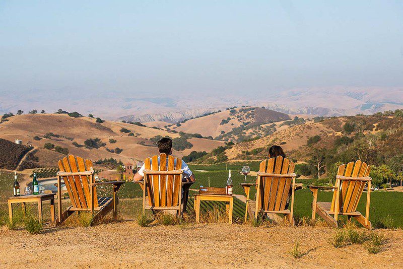 VisitUs-1000 Rear view of a man and a woman sitting in wooden Adirondack chairs overlooking a valley, with wine bottles and glasses on tables next to them