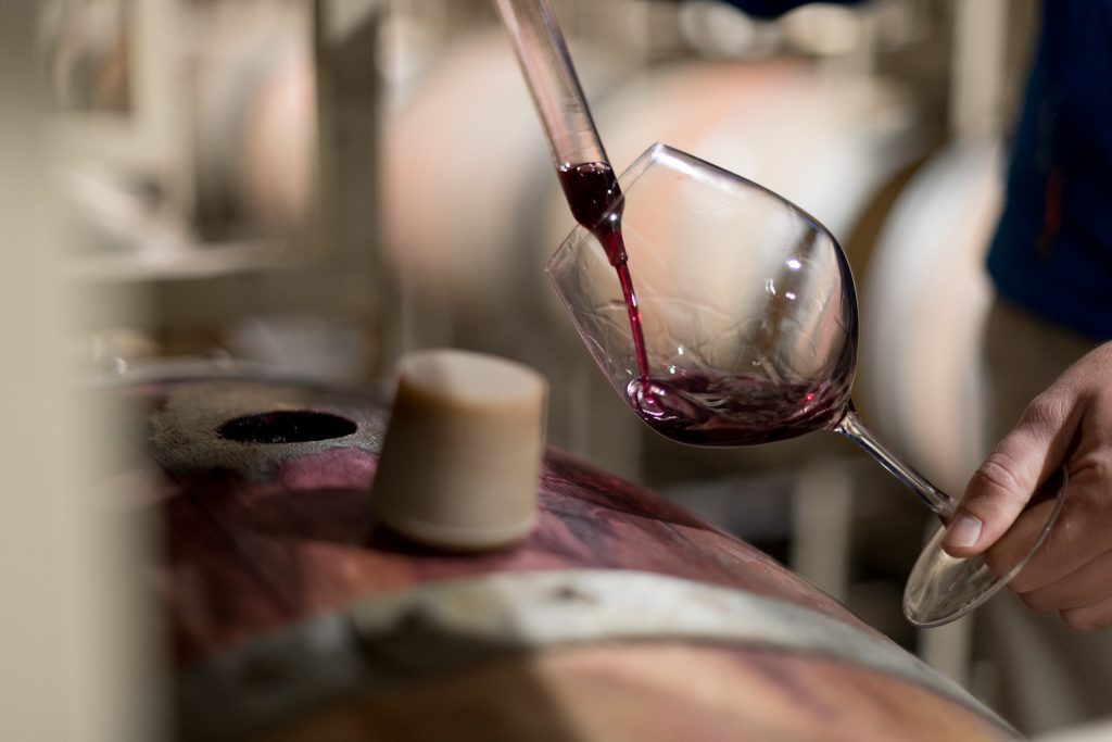 JointheClub A wine glass has red wine poured into it by a wine thief, next to an open barrel